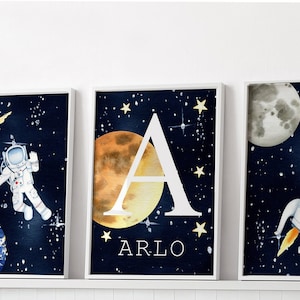 Puede incluir: Tres l&aacute;minas art&iacute;sticas enmarcadas con tem&aacute;tica espacial. Una presenta un astronauta, otra la letra "A" con el nombre "Arlo", y la tercera un cohete. Todas sobre un fondo azul oscuro estrellado.