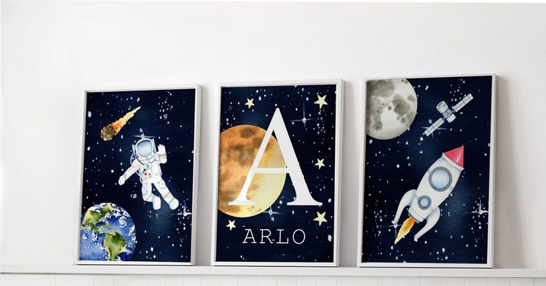 Puede incluir: Tres l&aacute;minas art&iacute;sticas enmarcadas con tem&aacute;tica espacial. Una presenta un astronauta, otra la letra "A" con el nombre "Arlo", y la tercera un cohete. Todas sobre un fondo azul oscuro estrellado.
