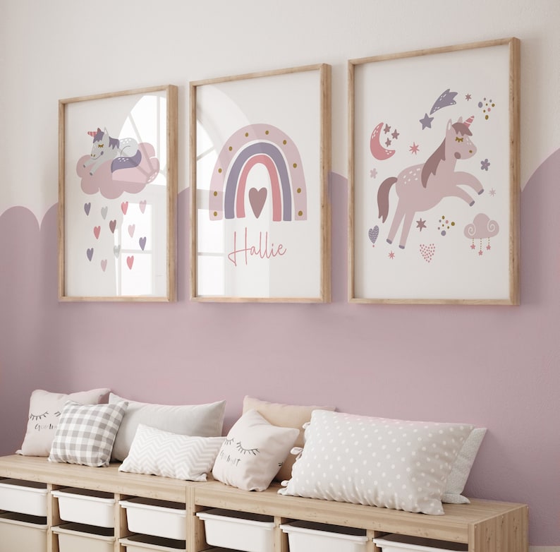 May include: Three framed art prints with a unicorn, rainbow, and stars theme. The prints feature soft pink, purple, and gold colors. One print has the name "Hallie" written in cursive. The prints are displayed above a wooden bench with pillows.