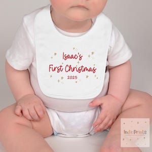 May include: White baby bib with the text "Isaac's First Christmas 2025" in red script, accented with gold stars. The bib is worn by a baby in a white onesie.