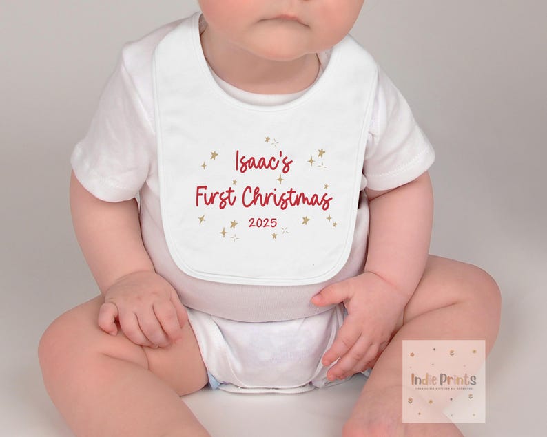 May include: White baby bib with the text "Isaac's First Christmas 2025" in red script, accented with gold stars. The bib is worn by a baby in a white onesie.