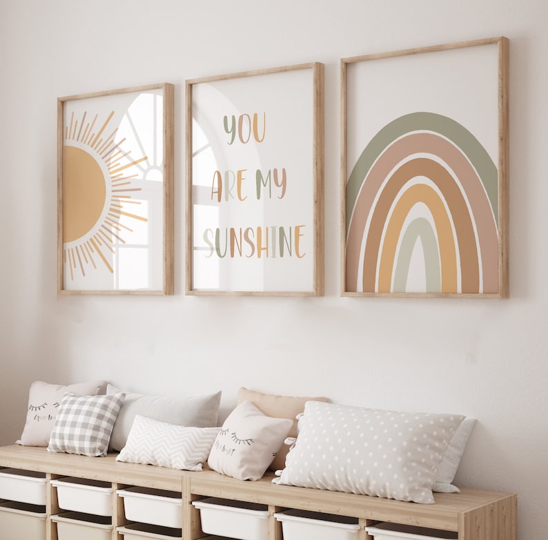 May include: Three framed art prints with a sun, a rainbow, and the text "YOU ARE MY SUNSHINE." The prints have a neutral color palette with shades of yellow, green, and brown. The frames are light wood. Below the prints are pillows and storage bins.
