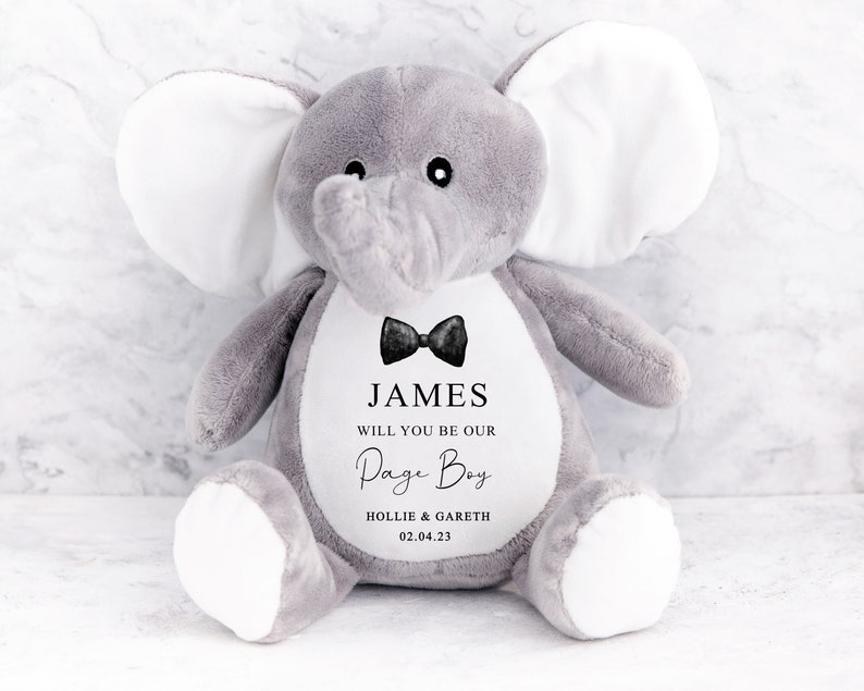 Pode incluir: Um brinquedo de pel&uacute;cia de elefante cinzento com grandes orelhas brancas. O elefante tem uma gravata borboleta preta e o texto "JAMES WILL YOU BE OUR Page Boy HOLLIE & GARETH 02.04.23" impresso no peito. Um presente fofo para um casamento.