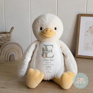 May include: A cream-colored plush duck toy with a yellow beak and feet. The belly features a printed design with a large grey letter E, a small rabbit illustration, and the text: Emmie Rae, born on 03/04/2026, weighing 5lb 7oz at 3.12am.
