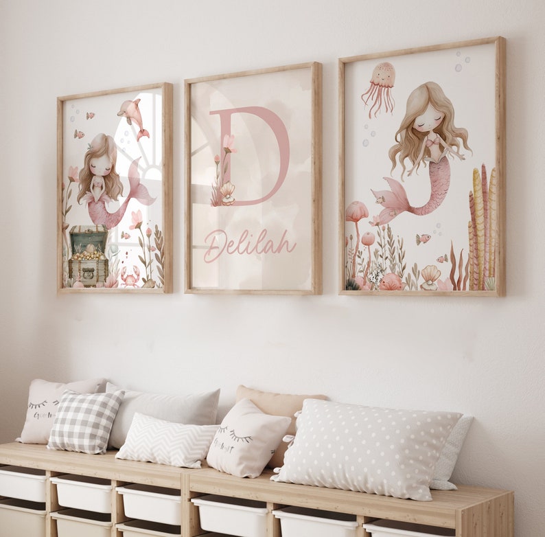 May include: Three framed mermaid art prints. The prints feature watercolor illustrations of mermaids, marine life, and the name "Delilah". The color palette includes pink, beige, and brown tones. The frames are made of light-colored wood.