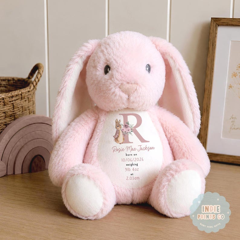 May include: A soft, plush light pink stuffed rabbit with long ears and a white belly patch. The belly features a printed design with the letter R, a rabbit illustration, and the text: Rosie Mae Jackson, born on 10/06/2026, weighing 9lb 4oz at 2.03am.