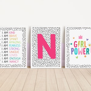May include: Three framed prints with white backgrounds and black polka dots. One print has the text "I AM KIND, BRAVE, SMART, AMAZING, FEARLESS, STRONG, UNIQUE, LOVED, ENOUGH, NANCY" in rainbow colors. Another features a large pink letter "N". The third says "GIRL POWER".