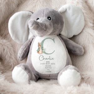 Puede incluir: Un peluche de elefante gris con detalles blancos en las orejas y los pies. El elefante tiene un dise&ntilde;o personalizado en su vientre con el nombre "Charlie", fecha de nacimiento, peso y hora. Tambi&eacute;n hay un conejito y un dise&ntilde;o floral.