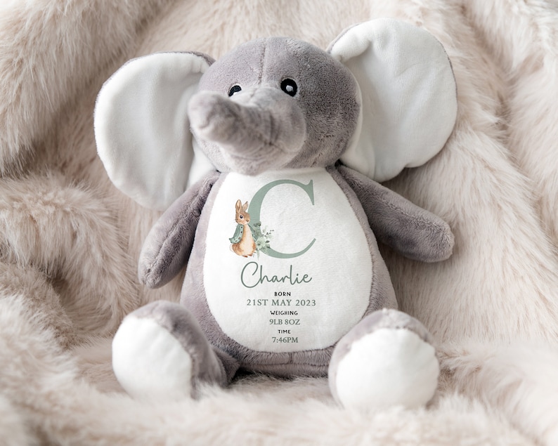 Puede incluir: Un peluche de elefante gris con detalles blancos en las orejas y los pies. El elefante tiene un dise&ntilde;o personalizado en su vientre con el nombre "Charlie", fecha de nacimiento, peso y hora. Tambi&eacute;n hay un conejito y un dise&ntilde;o floral.