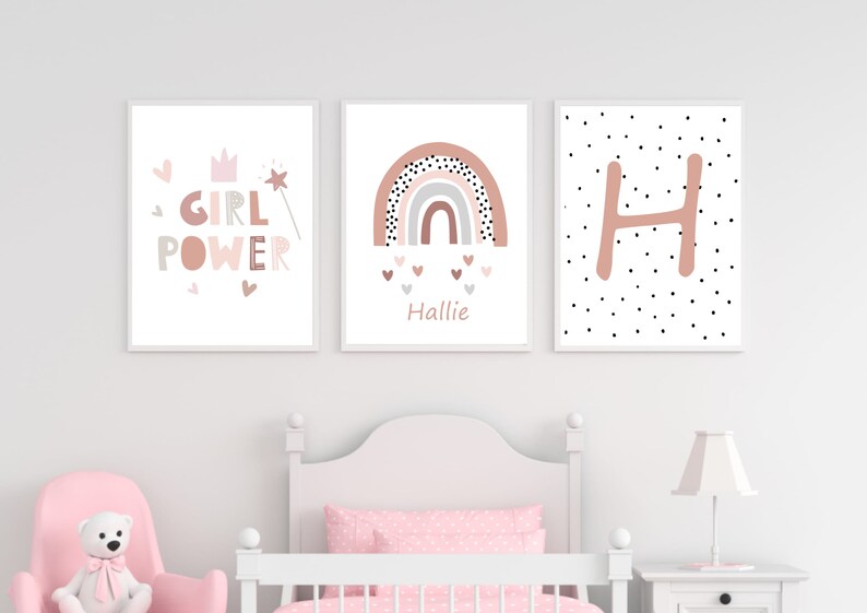 May include: Three framed art prints with a nursery theme. The prints feature the words "GIRL POWER", a rainbow design, and the letter "H". The color palette includes pink, gray, and white, creating a soft and inviting aesthetic.