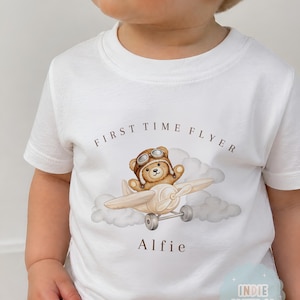 Könnte beinhalten: Ein weißes Kurzarm-T-Shirt mit einer Grafik eines braunen Teddybären, der eine Fliegerbrille trägt und ein kleines Flugzeug durch Wolken steuert. Der Text "FIRST TIME FLYER" steht über dem Bären und der Name "Alfie" ist darunter gedruckt.