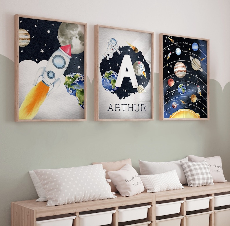 May include: Three framed watercolor prints with a space theme. One features a rocket ship, another has the letter "A" and the name "ARTHUR", and the third depicts the planets in our solar system. The prints are in wooden frames and are displayed above a wooden bench.