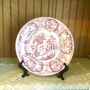 May include: A round ceramic plate featuring a maroon and white transferware pattern. The center depicts a landscape with buildings and palm trees, while the border is decorated with alternating floral panels and small architectural scenes.
