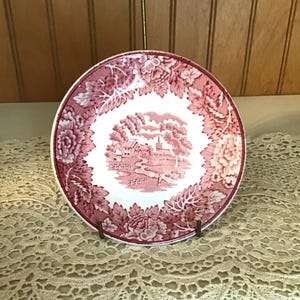 May include: A round ceramic saucer featuring a white center with a detailed red transferware landscape illustration of a village, trees, and a path. The wide border is decorated with a dense pattern of red floral motifs and leaves.
