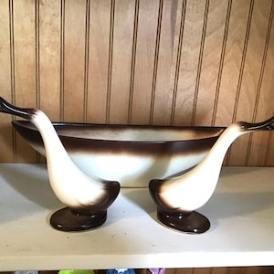 May include: A vintage ceramic centerpiece featuring a long, oval bowl with a brown and white gradient. Two swan-shaped candle holders, with white necks and brown bases, flank the bowl. The set is displayed on a white shelf against a wood-paneled wall.