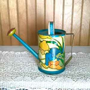 May include: Vintage metal watering can with a blue spout and handle. The can features a colorful illustration of a child in a garden. The can is painted with yellow, blue, and orange accents, and has a silver handle.