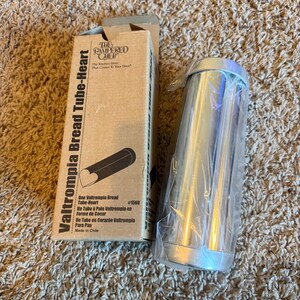 May include: A new Valtrompia Bread Tube-Heart baking mold from The Pampered Chef. The box is tan with black text and a drawing of the bread mold. The mold is silver and cylindrical, wrapped in clear plastic.