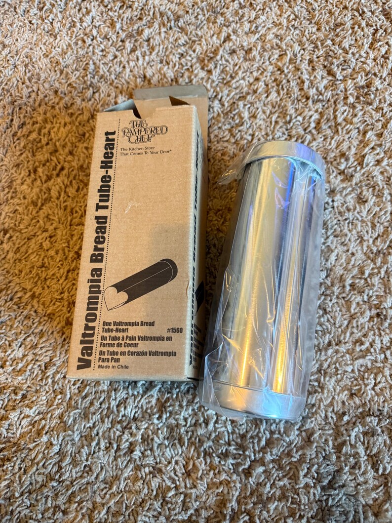 May include: A new Valtrompia Bread Tube-Heart baking mold from The Pampered Chef. The box is tan with black text and a drawing of the bread mold. The mold is silver and cylindrical, wrapped in clear plastic.
