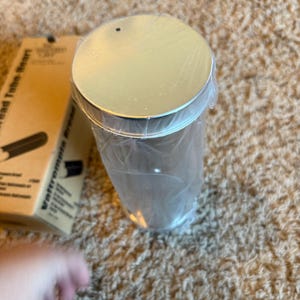 May include: A cylindrical metal container with a silver lid, wrapped in clear plastic. The container is on a textured, light brown surface. A cardboard box with text is partially visible in the background.