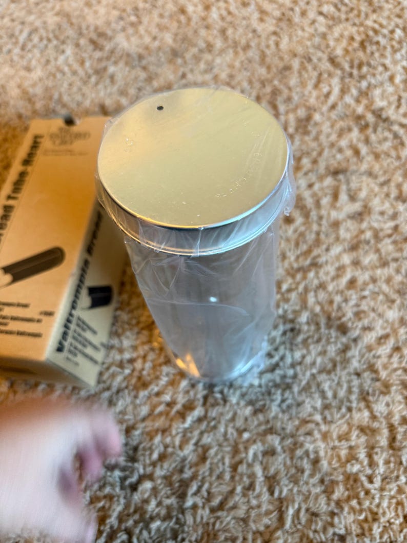 May include: A cylindrical metal container with a silver lid, wrapped in clear plastic. The container is on a textured, light brown surface. A cardboard box with text is partially visible in the background.