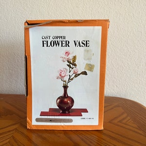 May include: A box for a cast copper flower vase. The box is orange and features a picture of a vase with pink roses. The text on the box reads "Cast Copper Flower Vase" and "CODE: 11-95-10".