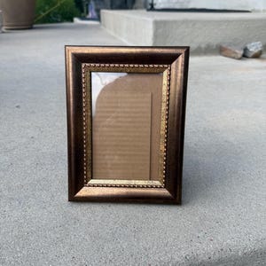 May include: A rectangular picture frame with a dark bronze finish and a gold-toned inner border featuring a beaded texture. The frame has a clear glass front and a plain brown cardboard insert visible inside the display area of the frame.