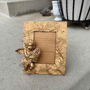May include: A gold-colored picture frame with a textured surface and a cherub figurine holding flowers. The frame has a rectangular opening for a photo and is placed on a concrete surface.