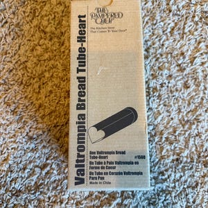 May include: A cardboard box for a Valtrompia Bread Tube-Heart. The box is tan with black text and a black illustration of the tube with a heart-shaped end. The text includes "The Pampered Chef" and "The Kitchen Store That Comes To Your Door."