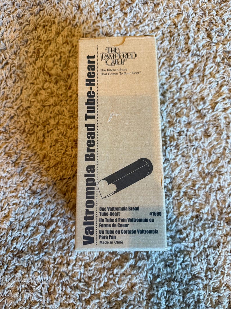 May include: A cardboard box for a Valtrompia Bread Tube-Heart. The box is tan with black text and a black illustration of the tube with a heart-shaped end. The text includes "The Pampered Chef" and "The Kitchen Store That Comes To Your Door."