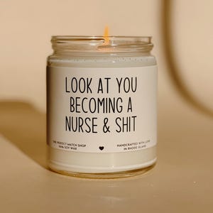 May include: A lit, white soy wax candle in a glass jar. The label reads "LOOK AT YOU BECOMING A NURSE & SHIT." Additional text includes "THE PERFECT MATCH SHOP" and "HANDCRAFTED WITH LOVE IN RHODE ISLAND."