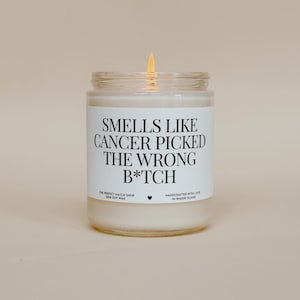 Puede incluir: Una vela encendida en un tarro de vidrio transparente con una etiqueta blanca. La etiqueta dice "SMELLS LIKE CANCER PICKED THE WRONG B*TCH". El texto "THE PERFECT MATCH SHOP 100% SOY WAX" y "HANDCRAFTED WITH LOVE IN RHODE ISLAND" también están impresos.