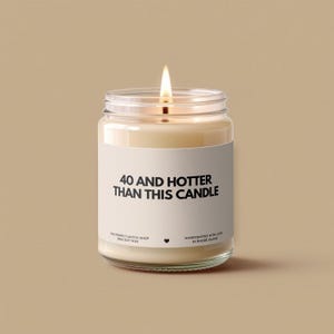 May include: A lit soy wax candle in a clear glass jar. The candle has a white label with the text "40 AND HOTTER THAN THIS CANDLE". The candle is handcrafted with love in Rhode Island.