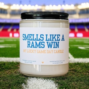 Puede incluir: Una vela en tarro de cristal con tapa negra, con una etiqueta blanca con texto azul que dice "SMELLS LIKE A RAMS WIN" y "MY LUCKY GAME DAY CANDLE". La vela está sobre un campo verde.