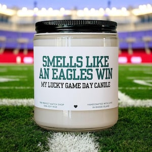 Op de afbeelding: Een glazen potkaars met een zwart deksel, met het opschrift "Smells Like An Eagles Win, My Lucky Game Day Candle". Het etiket bevat ook tekst over de winkel en sojawas. De kaars staat op een groen veld.