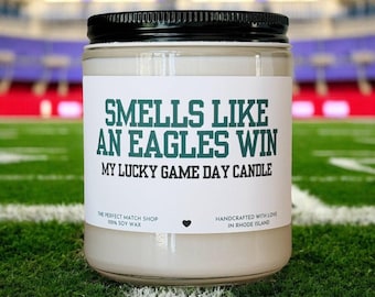 Könnte beinhalten: Ein Glas-Kerzenglas mit schwarzem Deckel, beschriftet mit "Smells Like An Eagles Win, My Lucky Game Day Candle". Das Etikett enthält auch Text über den Shop und Sojawachs. Die Kerze steht auf einem grünen Feld.