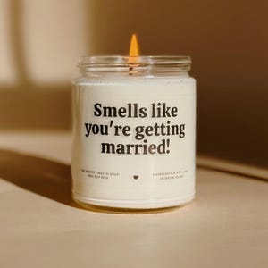 May include: A burning candle in a glass jar with the text "Smells like you're getting married!". The candle is white and from The Perfect Match Shop, made with 100% soy wax. It is handcrafted in Rhode Island.