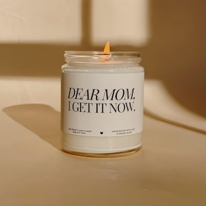 Könnte beinhalten: Eine brennende weiße Kerze in einem durchsichtigen Glas. Auf dem Etikett der Kerze steht "DEAR MOM, I GET IT NOW". Das Etikett enthält auch den Text "THE PERFECT MATCH SHOP 100% SOY WAX" und "HANDCRAFTED WITH LOVE IN RHODE ISLAND."