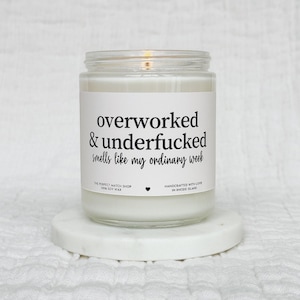 May include: A glass jar candle with a white label, reading "overworked & underfucked smells like my ordinary week." The candle is on a white marble coaster. The text "THE PERFECT MATCH SHOP 100% SOY WAX" is also visible. The candle is lit.