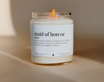 Könnte beinhalten: Eine brennende Sojawachskerze in einem durchsichtigen Glas. Das Etikett trägt die Aufschrift "maid of honor" mit einer Definition. Die Kerze wird in Rhode Island handgefertigt.