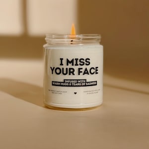 May include: A lit white soy wax candle in a glass jar. The candle has the text "I MISS YOUR FACE" in black, with additional text below. The candle is infused with warm hugs and tears of sadness.