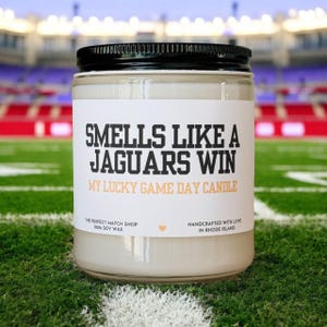 May include: A clear glass jar candle with a black lid, labeled "SMELLS LIKE A JAGUARS WIN." The text "MY LUCKY GAME DAY CANDLE" is below. The candle is on a green field with white lines, with a stadium in the background.