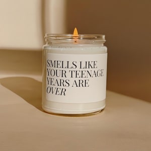 Op de afbeelding: Een brandende kaars in een heldere glazen pot met een wit etiket. Het etiket toont de tekst "Smells Like Your Teenage Years Are Over" in zwarte letters. De kaars geeft een warm, uitnodigend licht.