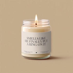 May include: A lit, cream-coloured soy wax candle in a clear glass jar. The label reads "SMELLS LIKE HE FINALLY PUT A RING ON IT." The candle is handcrafted in Rhode Island. The jar is approximately 7.6 cm tall.