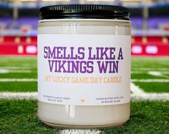May include: A glass jar candle with a black lid, labeled "SMELLS LIKE A VIKINGS WIN." The text "MY LUCKY GAME DAY CANDLE" is below the main text. The candle is on a green surface with white lines, resembling a football field.