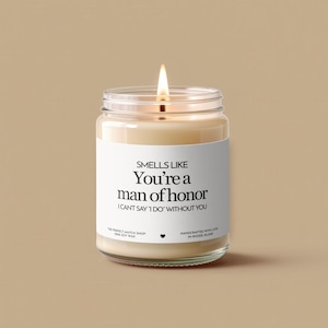 Może przedstawiać: Zapalone świeca sojowa w przezroczystym szklanym słoiku z białą etykietą. Na etykiecie widnieje napis "Smells Like You're a man of honor I can't say I do without you." Świeca jest ręcznie robiona na Rhode Island.