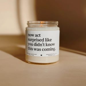 Könnte beinhalten: Ein Glas-Kerzenglas mit einem weißen Etikett mit dem Text "now act surprised like you didn't know this was coming." Die Kerze ist mit cremefarbenem Wachs gefüllt und hat einen Metalldeckel.