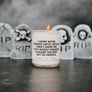 Puede incluir: Una vela blanca encendida en un tarro de vidrio, con un mensaje humorístico en la etiqueta, sobre un fondo de lápidas grises. La etiqueta de la vela dice: "Espero que seamos amigos hasta que muramos, luego espero que sigamos siendo amigos fantasmas y asustemos a la gente."