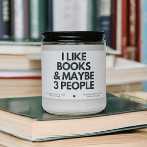 Puede incluir: Un tarro de vela blanco con tapa negra, con el texto "I LIKE BOOKS & MAYBE 3 PEOPLE" en negro. La vela está colocada sobre una pila de libros, con más libros en el fondo. La vela está hecha con cera de soja 100%.