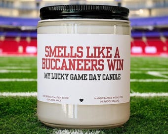 May include: A glass jar candle with a black lid, sitting on green grass. The label reads "SMELLS LIKE A BUCCANEERS WIN, MY LUCKY GAME DAY CANDLE." The candle is made with 100% soy wax and handcrafted in Rhode Island.
