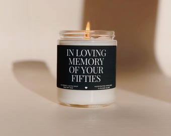 May include: A lit candle in a clear glass jar. The black label reads "IN LOVING MEMORY OF YOUR FIFTIES". The candle is made with soy wax. The jar is on a neutral background.